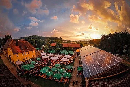 Luftbild eines großen Biergartens neben einer großen Scheune mit Solaranlage. Im Biergarten stehen viele Bierzelttische und Bänke aneinander gereiht. Darüber stehen grüne Sonnenschirme.